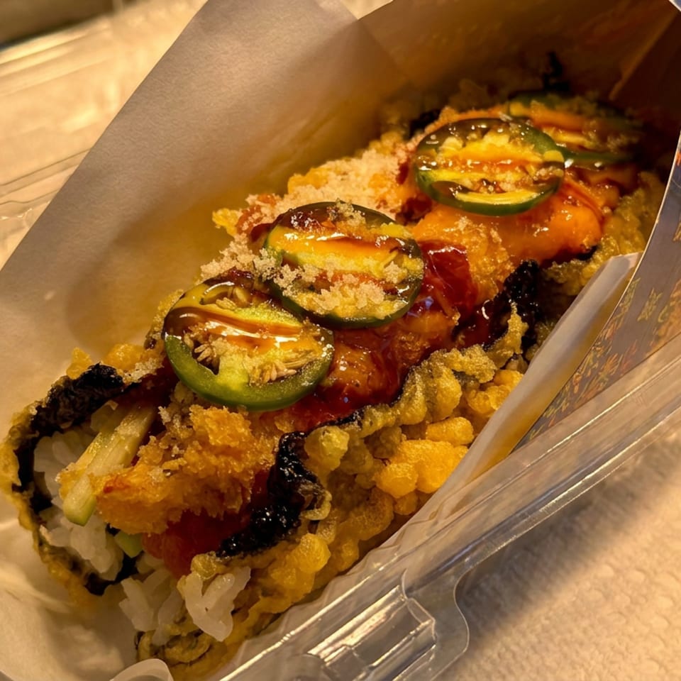 A crispy sushi roll in a takeout box, topped with sliced jalapeños, red sauce, and crunchy tempura bits.