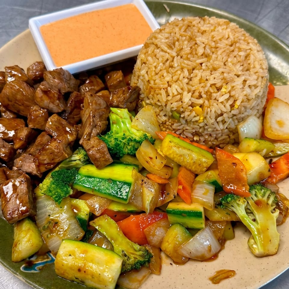 Plate of glazed beef cubes, fried rice, mixed sautéed vegetables, and orange dipping sauce on a restaurant table.