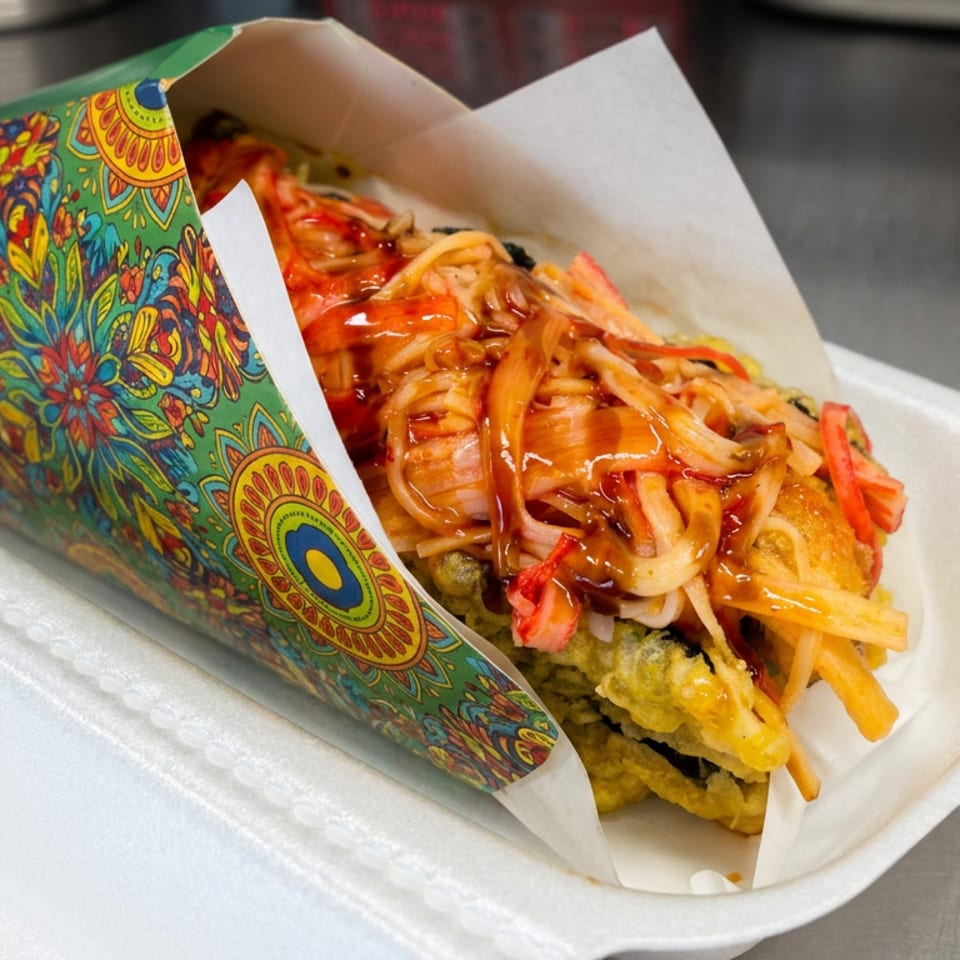 Crispy fried sandwich filled with shredded vegetables and red sauce in a colorful patterned paper wrapper on a white plate.