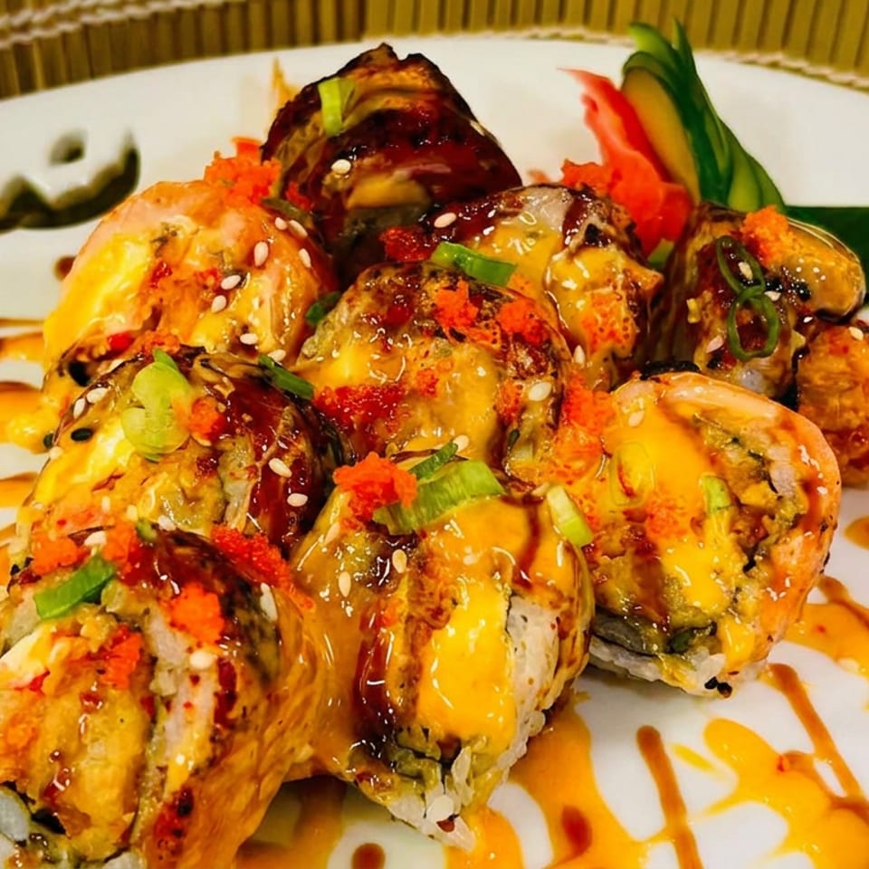 Sushi rolls topped with shrimp, spicy sauce, sesame seeds, and scallions, arranged with vegetable garnish on a white plate.